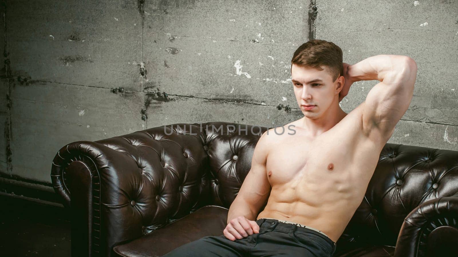 Young sexy men bodybuilder athlete sitting on a black leather couch with a naked torso and black trousers, Threw one's hands behind his head, studio portrait