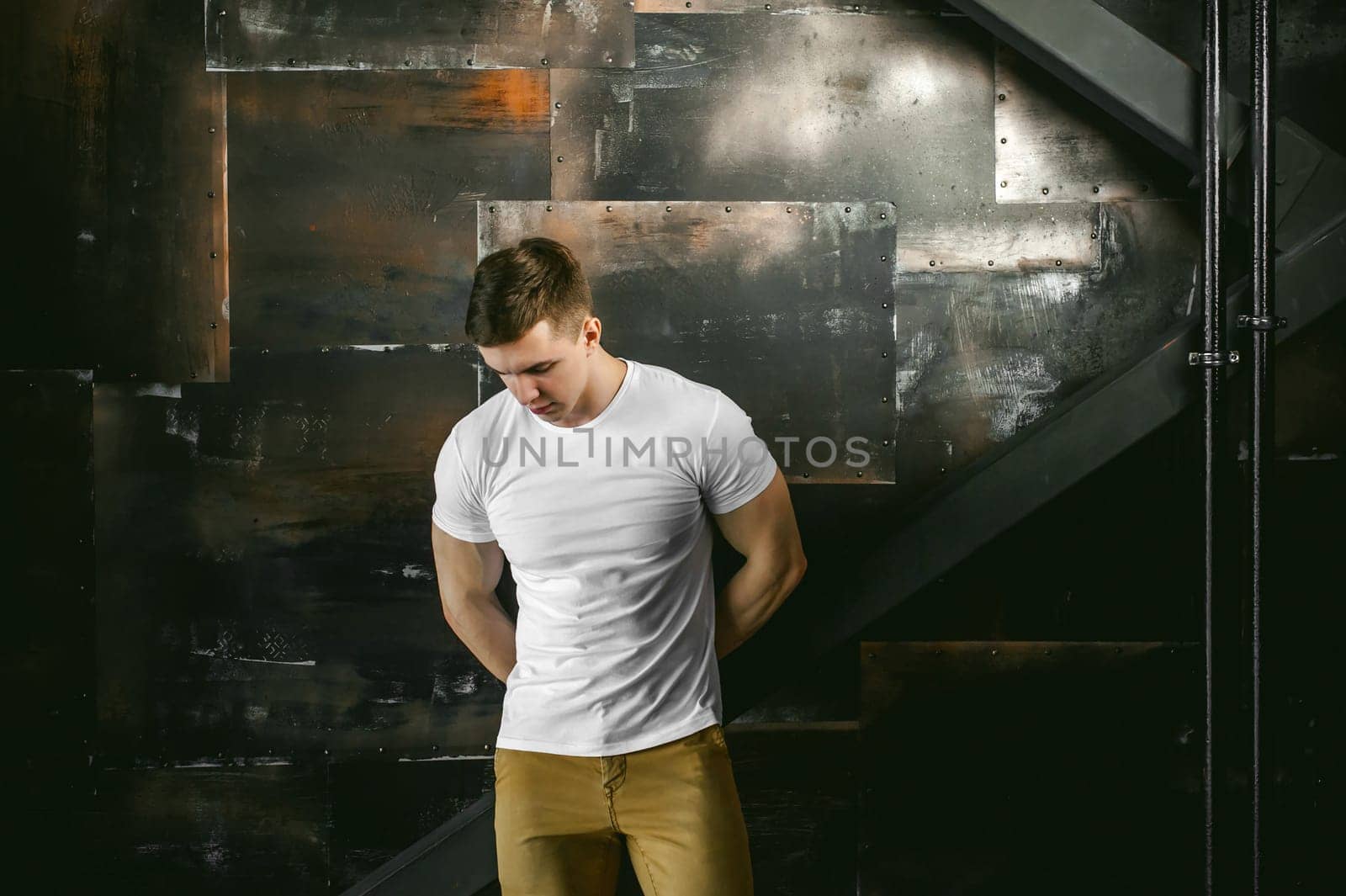 Young sexy men bodybuilder athlete, Studio portrait in a loft on the background of a stylized metal wall, a guy model in a white T-shirt and brown trousers