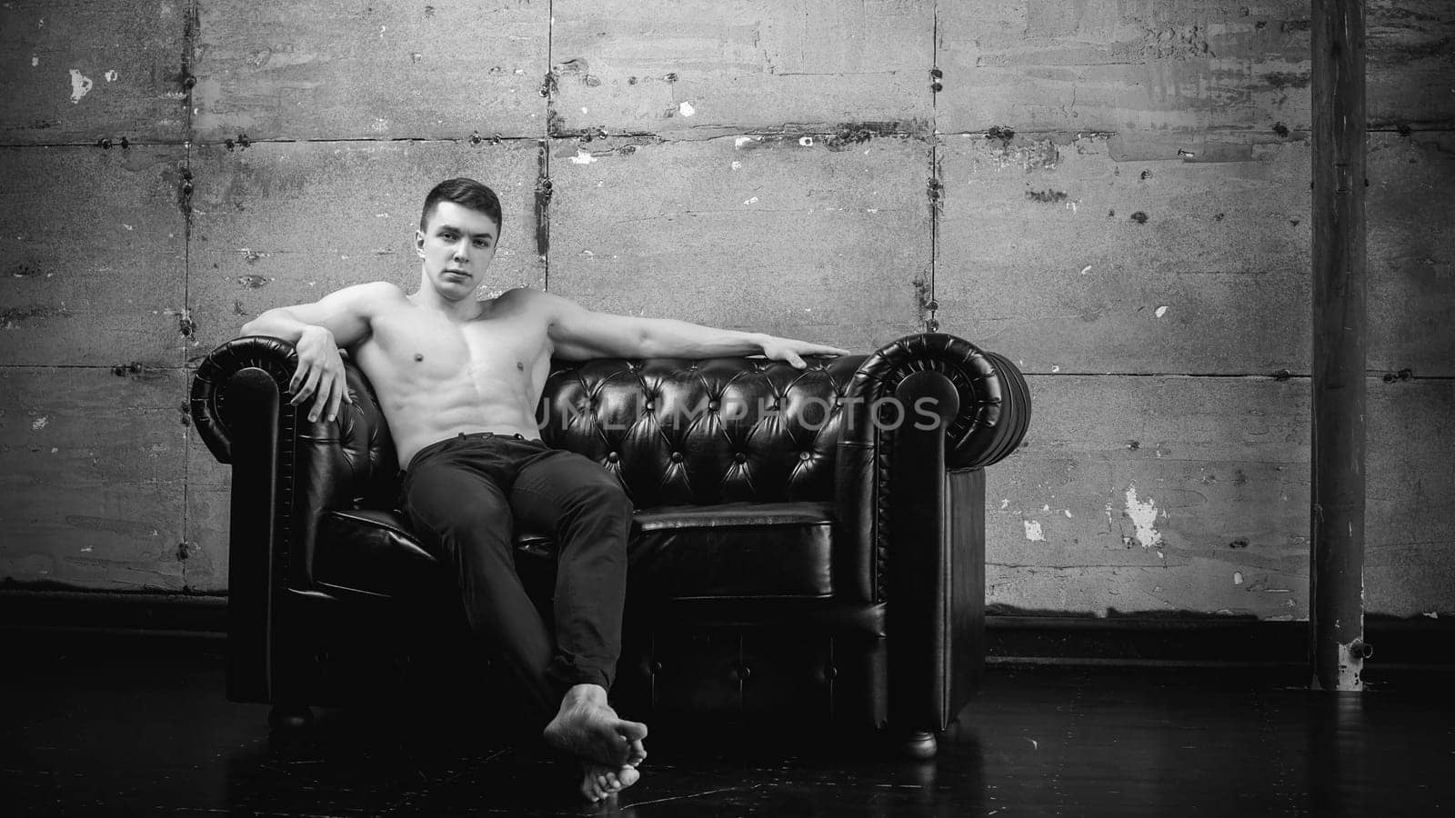 Young sexy men bodybuilder athlete sitting on a black leather couch with a naked torso and black trousers, studio portrait
