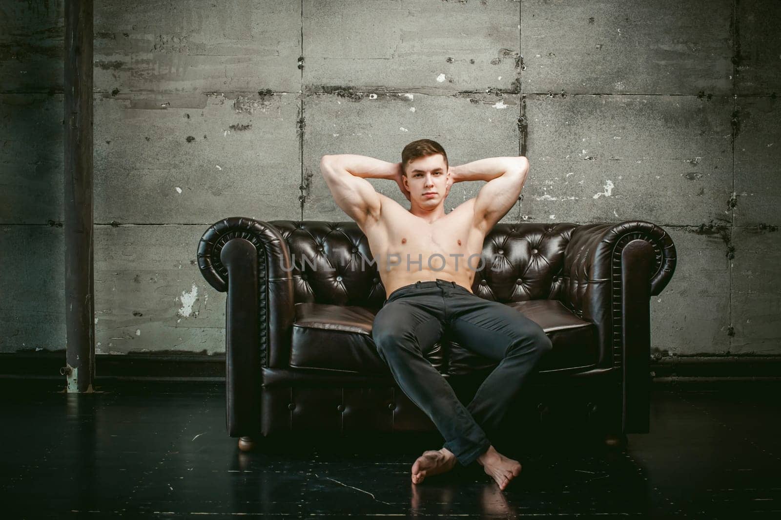 Young sexy men bodybuilder athlete sitting on a black leather couch with a naked torso and black trousers, Threw one's hands behind his head, studio portrait