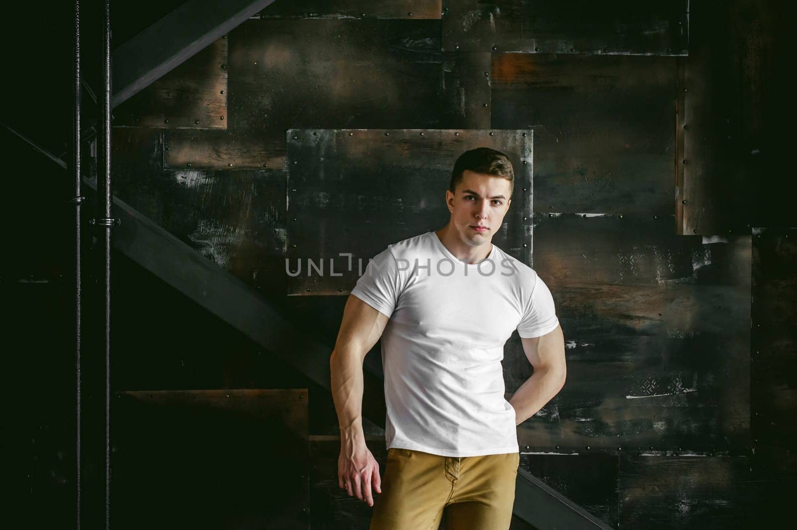 Young sexy men bodybuilder athlete, Studio portrait in a loft on the background of a stylized metal wall, a guy model in a white T-shirt and brown trousers