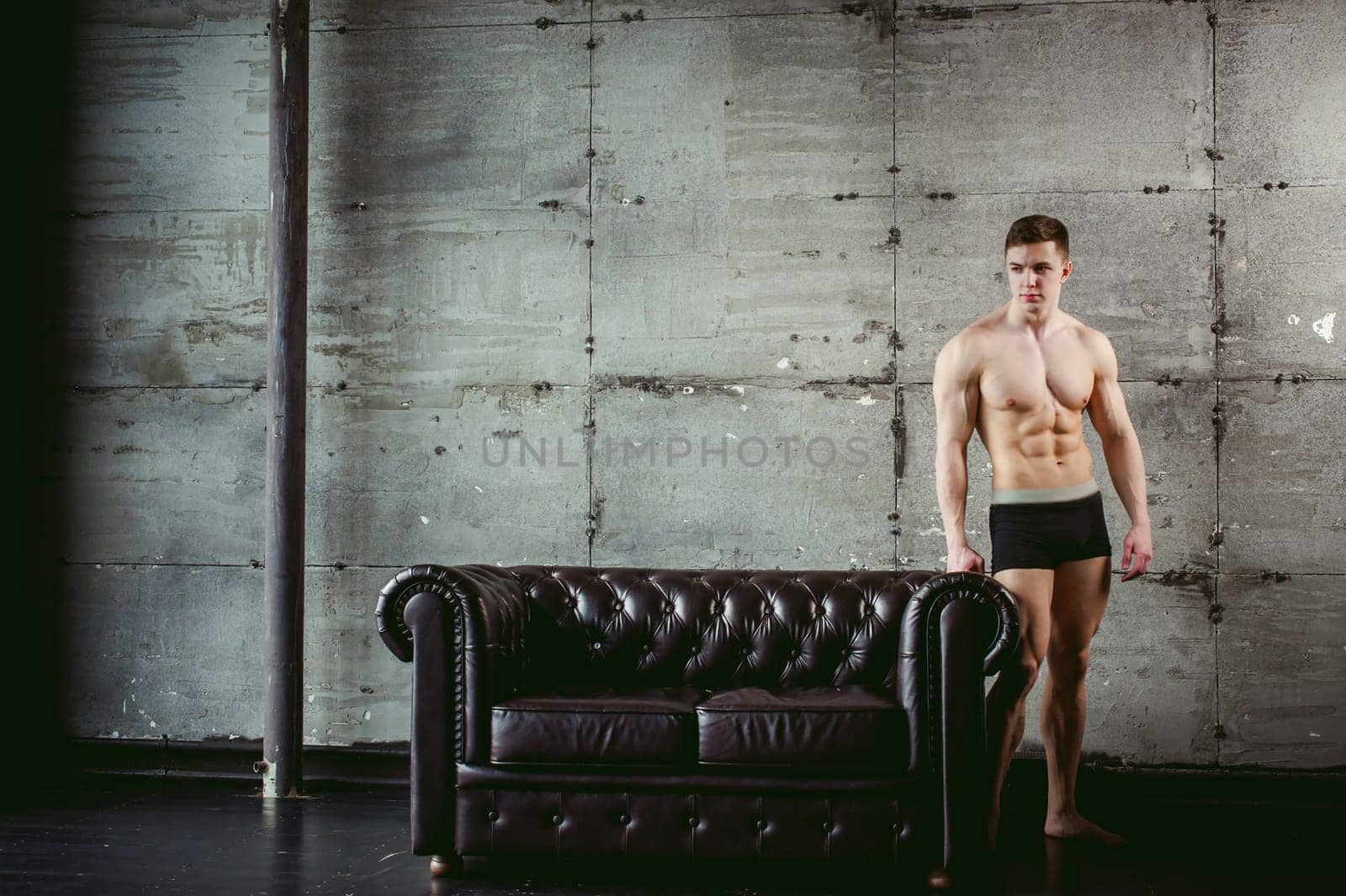 Young sexy men bodybuilder athlete stands at black leather couch with a naked torso, studio portrait