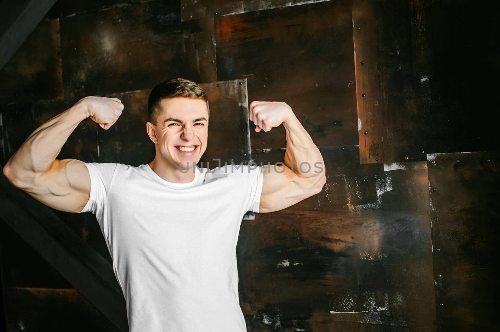 Young sexy men bodybuilder athlete, studio portrait in a loft on a background of  stylized metal wall, a guy model in a white T-shirt and brown trousers, tensed his arms showing muscles, bared teeth