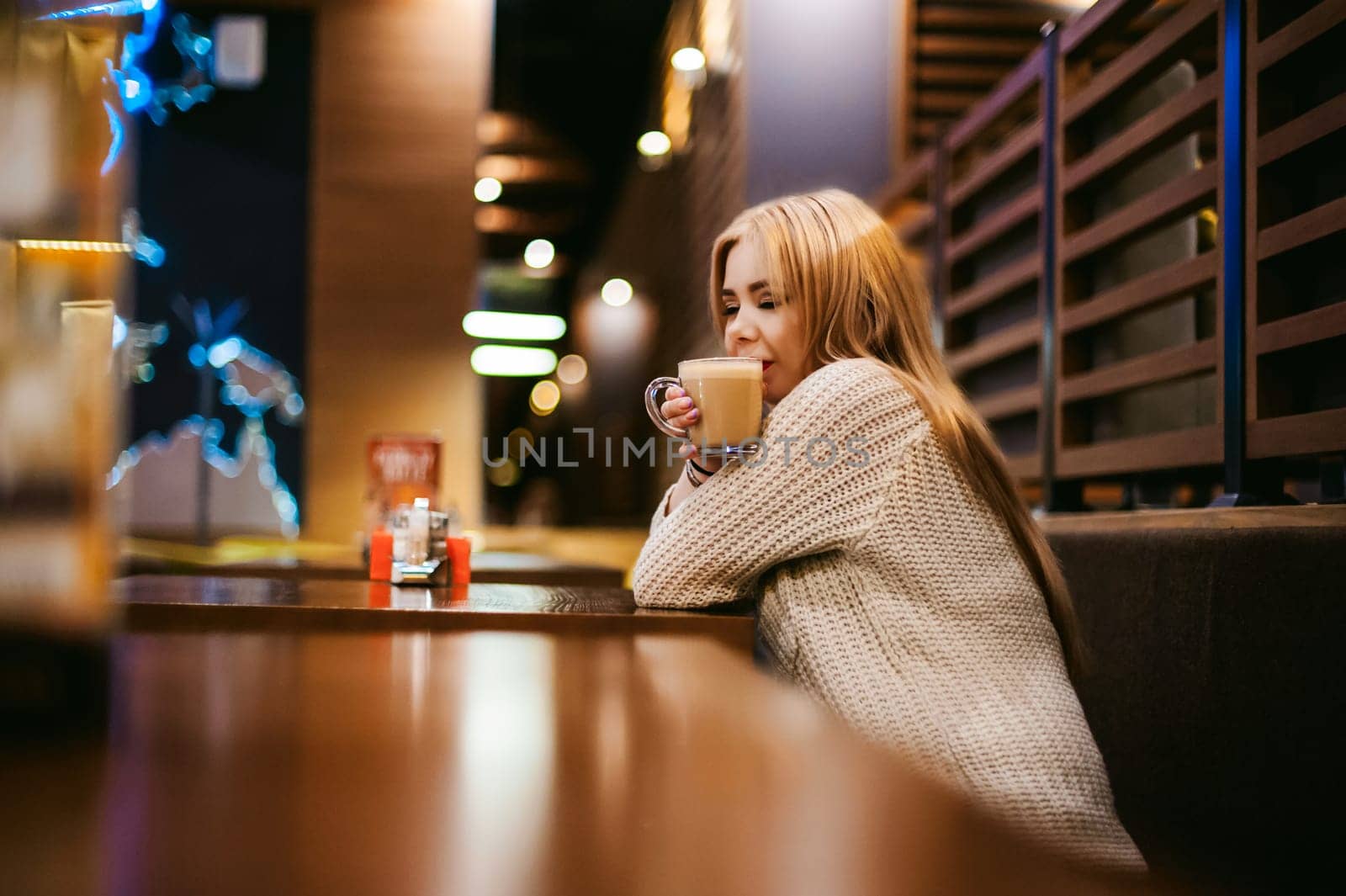 Cute young blonde woman drinks hot coffee from a glass cup in the interior of a coffee house