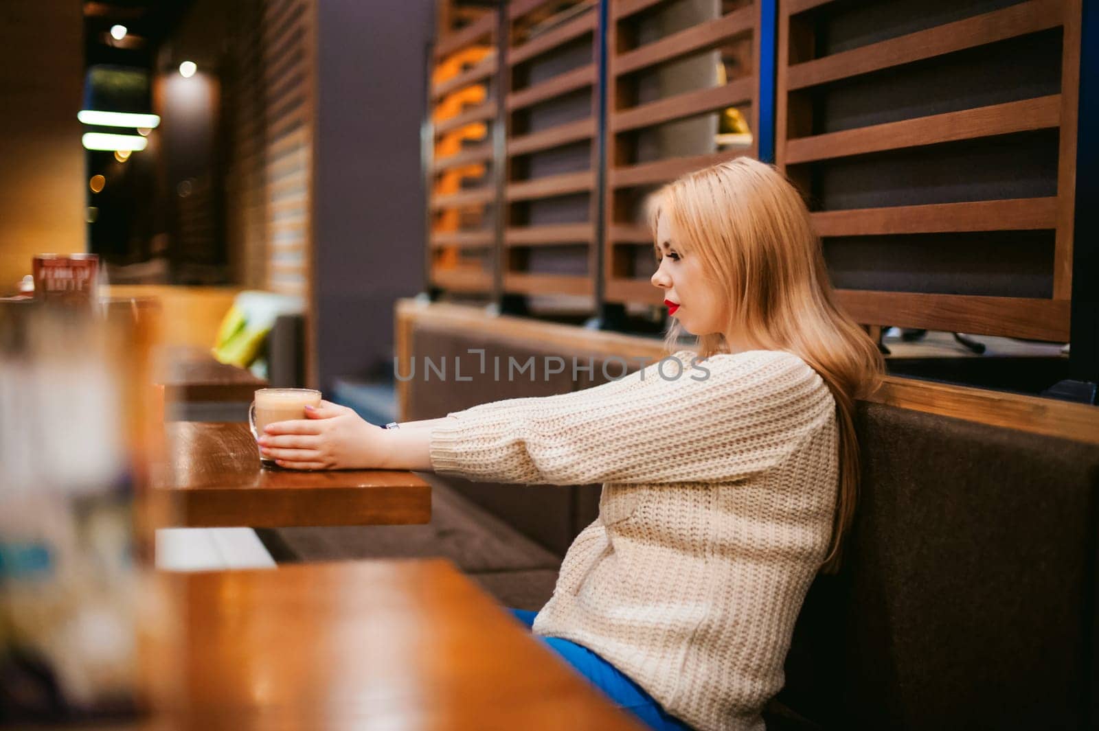 Cute young blonde woman drinks hot coffee from a glass cup in the interior of a coffee house