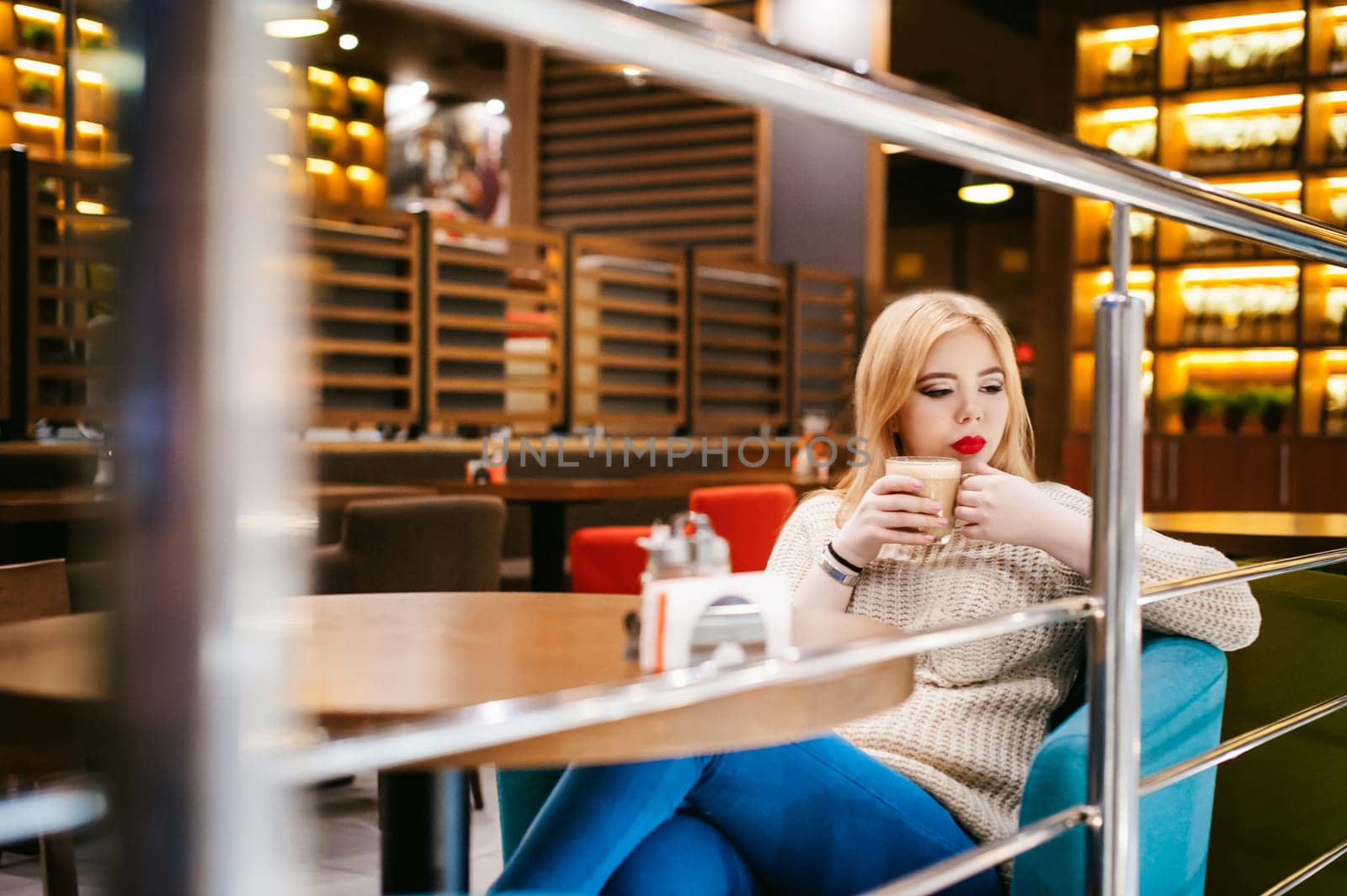 Cute young blonde woman drinks hot coffee from a glass cup in the interior of a coffee house