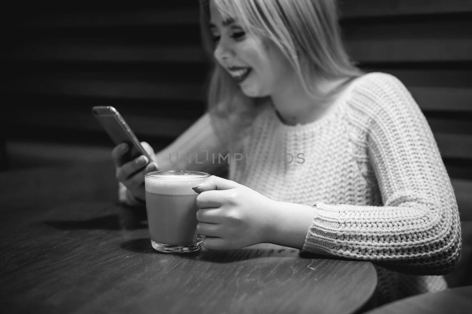 Young blonde woman in a knitted sweater sitting at a table in a cafe with a cup of coffee in her hands, spending time alone, doing internet communication through wifi