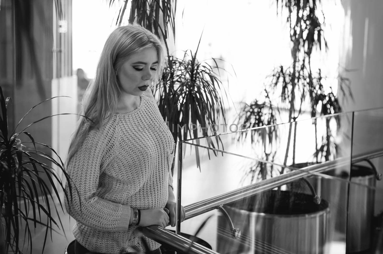 Portrait of a young woman dressed in a knitted sweater, standing near the handrails at a glass fence against the background of the office environment and indoor plant palm trees