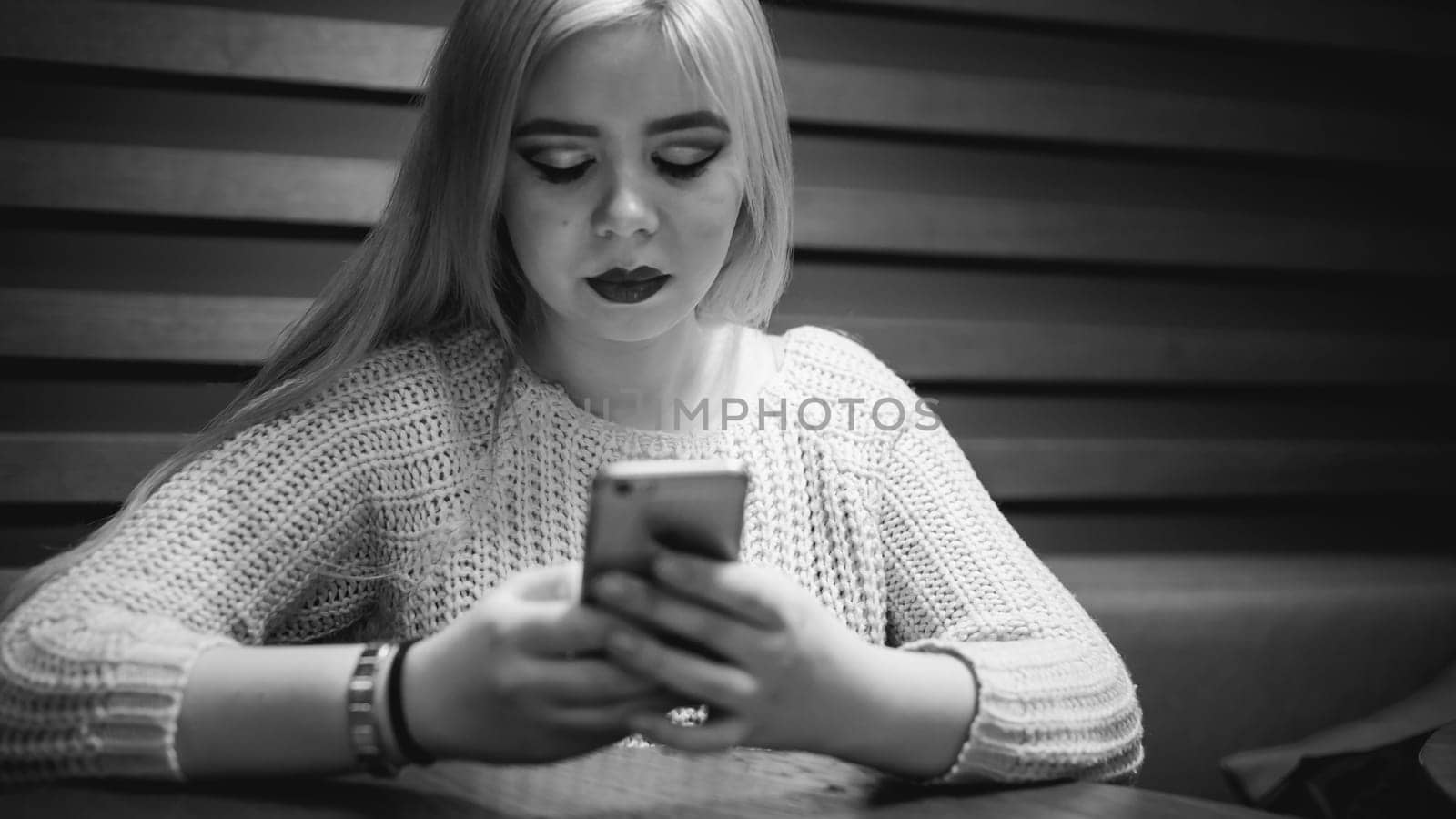 Young blond woman in a knitted sweater, sitting at a table in a cafe, spending time alone, doing internet communication through wifi