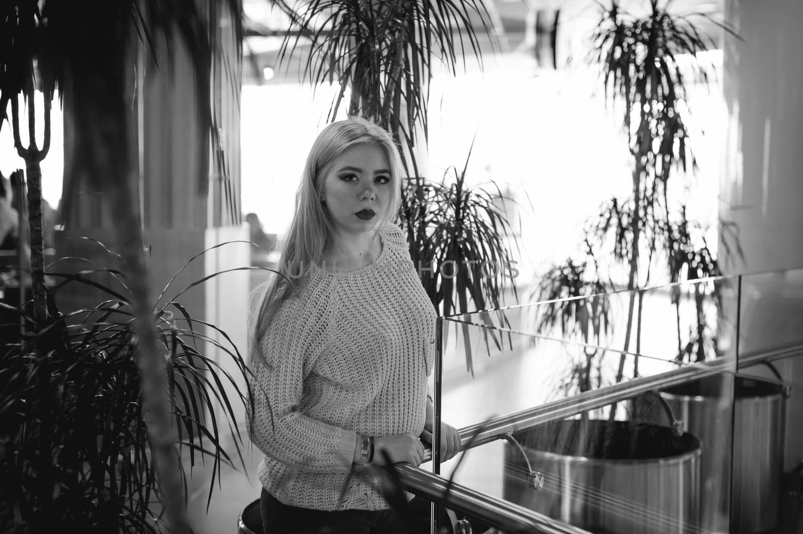 Portrait of a young woman dressed in a knitted sweater, standing near the handrails at a glass fence against the background of the office environment and indoor plant palm trees