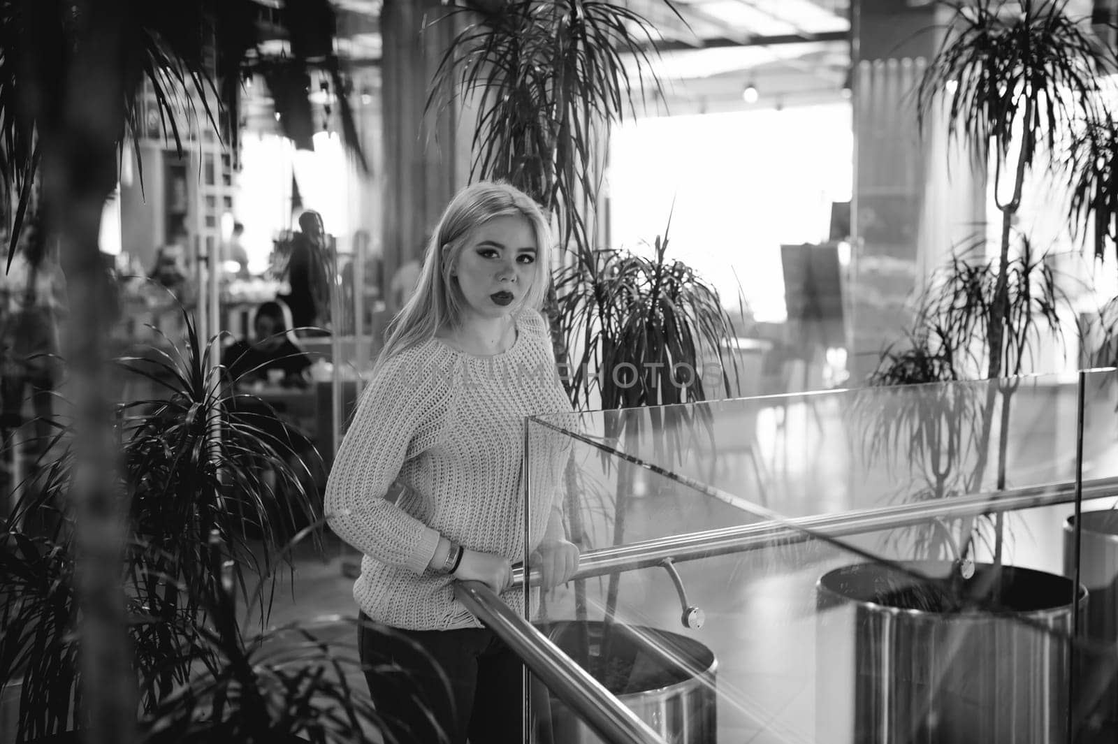 Portrait of a young woman dressed in a knitted sweater, standing near the handrails at a glass fence against the background of the office environment and indoor plant palm trees