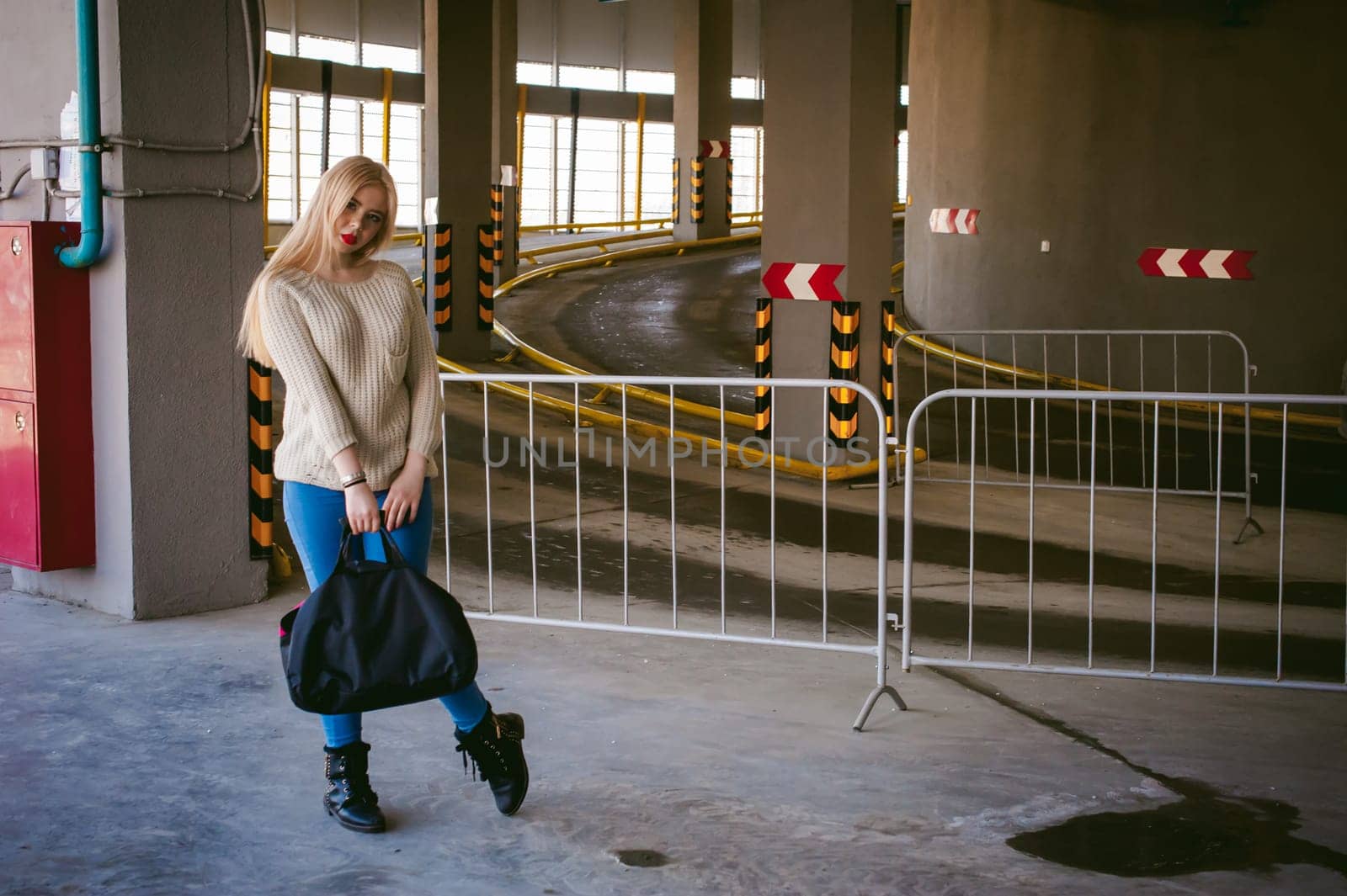 Portrait of a young woman dressed in a knitted sweater and blue jeans, holds in her hand a sports bag. standing on the background of a climb to the parking floor