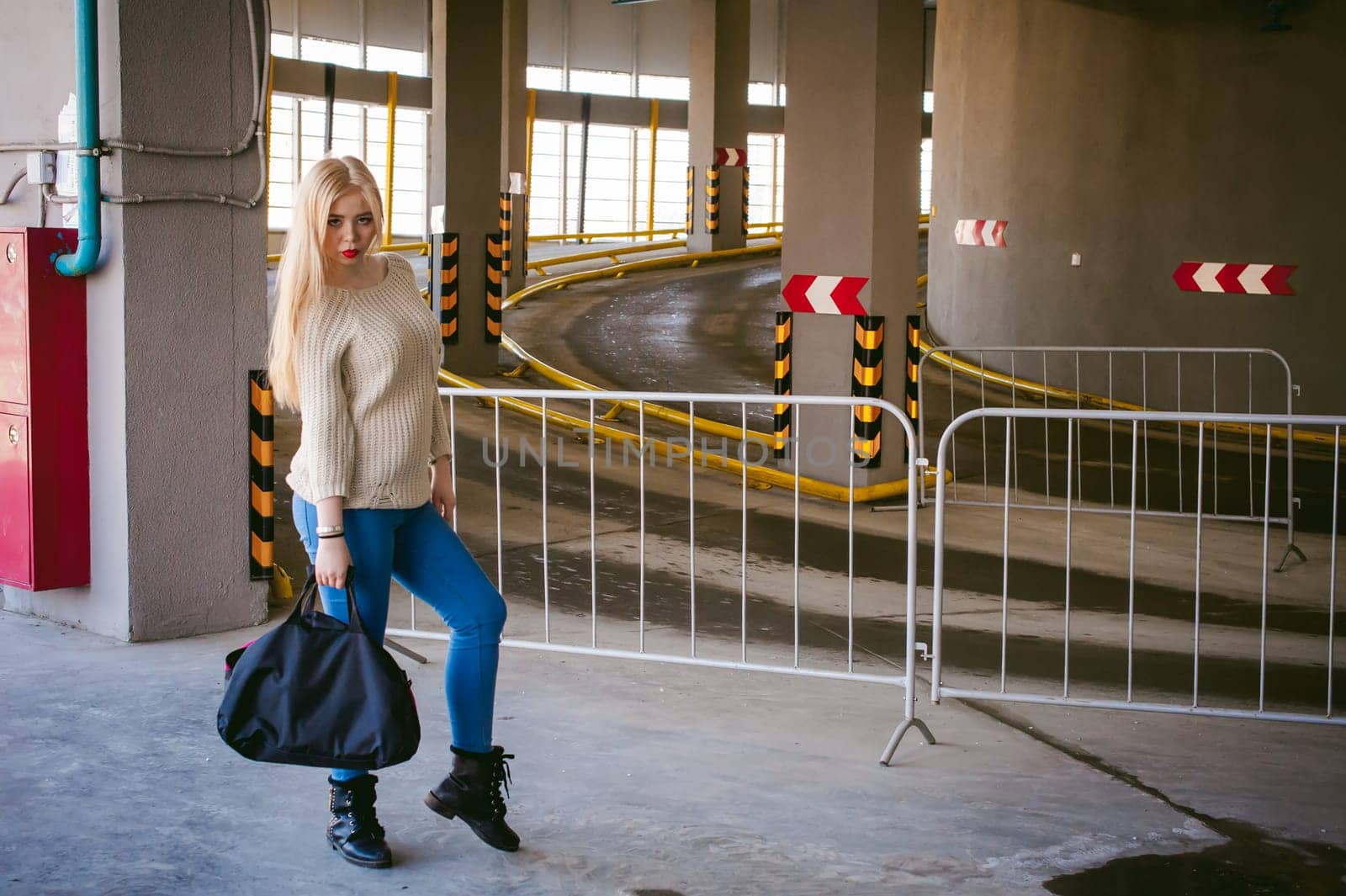 Portrait of a young woman dressed in a knitted sweater and blue jeans, holds in her hand a sports bag. standing on the background of a climb to the parking floor