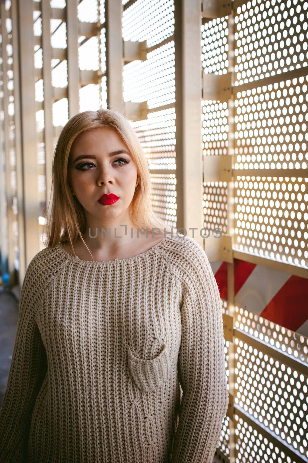 Portrait of a young woman dressed in a knitted sweater and blue jeans. Standing near a metal perforated parking fence construction