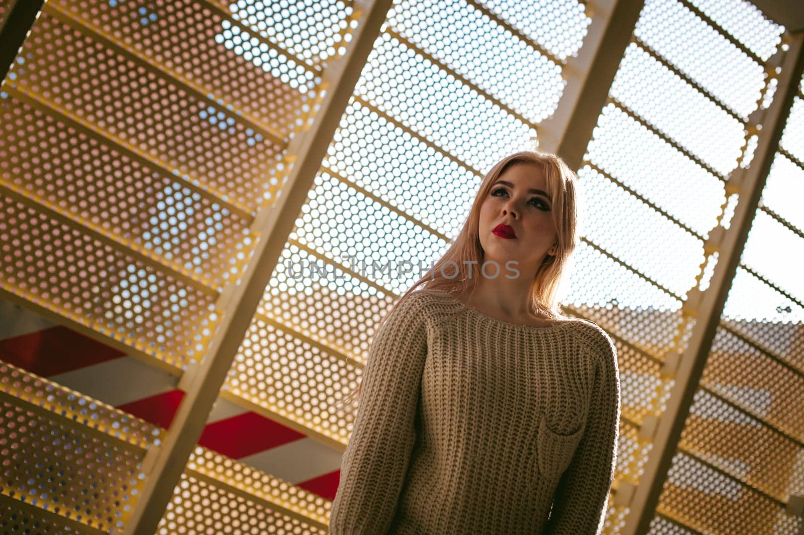 Portrait of a young woman dressed in a knitted sweater and blue jeans. Standing near a metal perforated parking fence construction