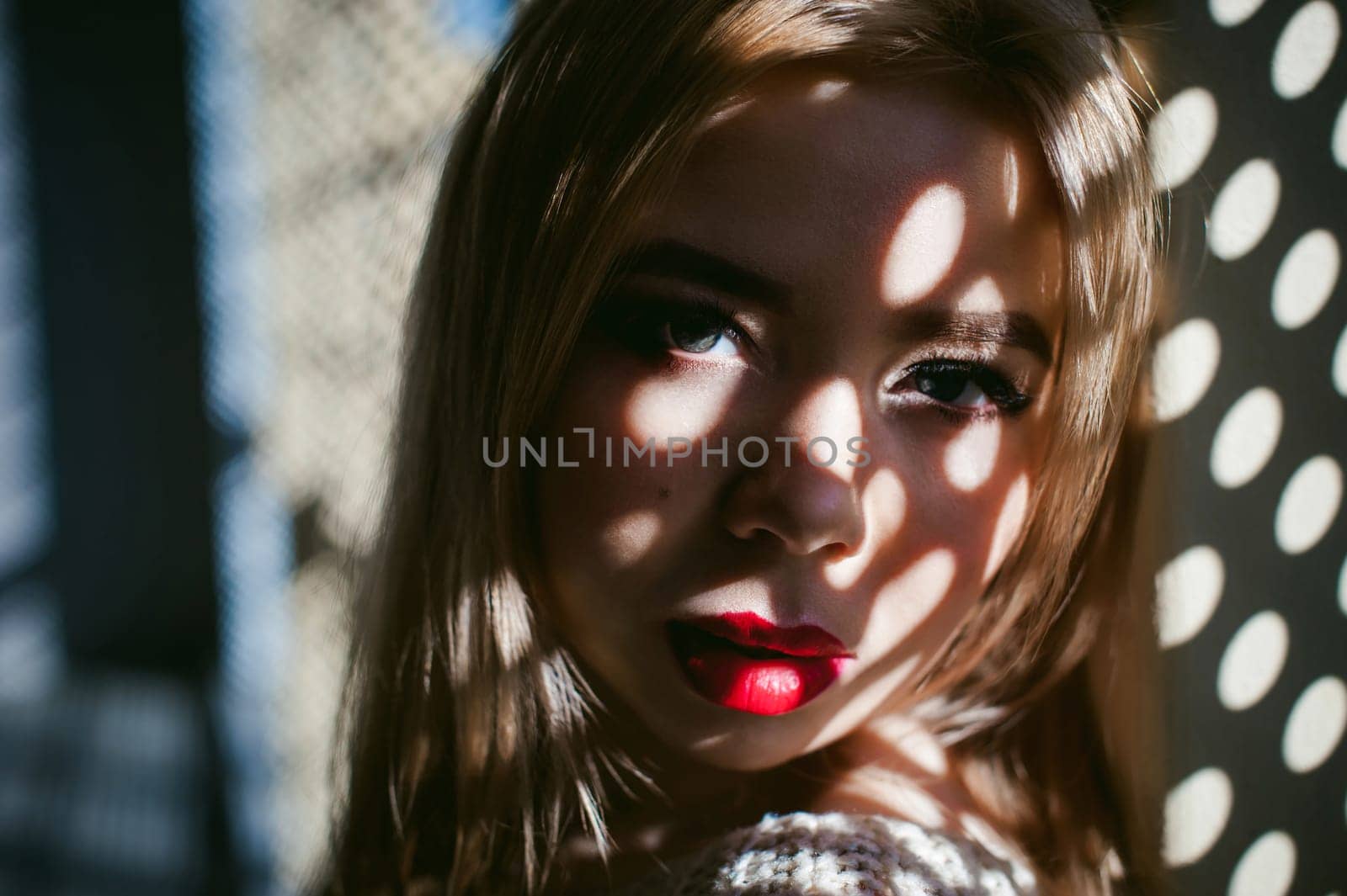 Portrait of a young woman dressed in a knitted sweater and blue jeans. Standing near a metal perforated parking fence construction. Shadows in the form of dots of light fall on her face