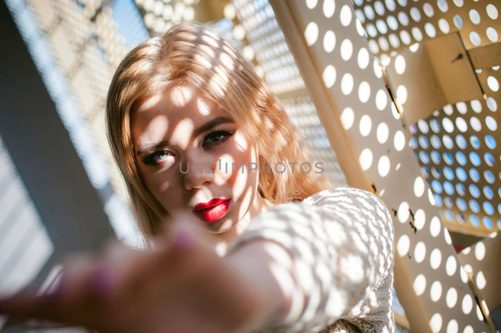 Portrait of a young woman dressed in a knitted sweater and blue jeans. Standing near a metal perforated parking fence construction. Shadows in the form of dots of light fall on her face