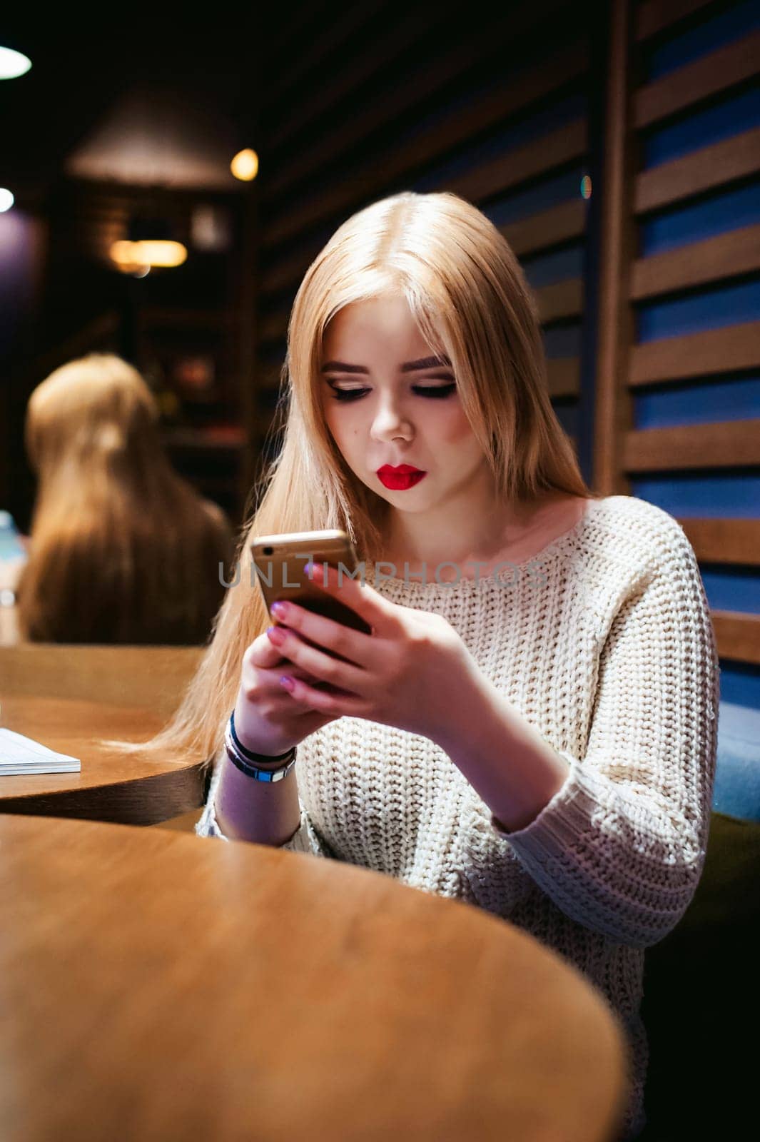 Young blond woman in a knitted sweater, sitting at a table in a cafe, spending time alone, doing internet communication through wifi