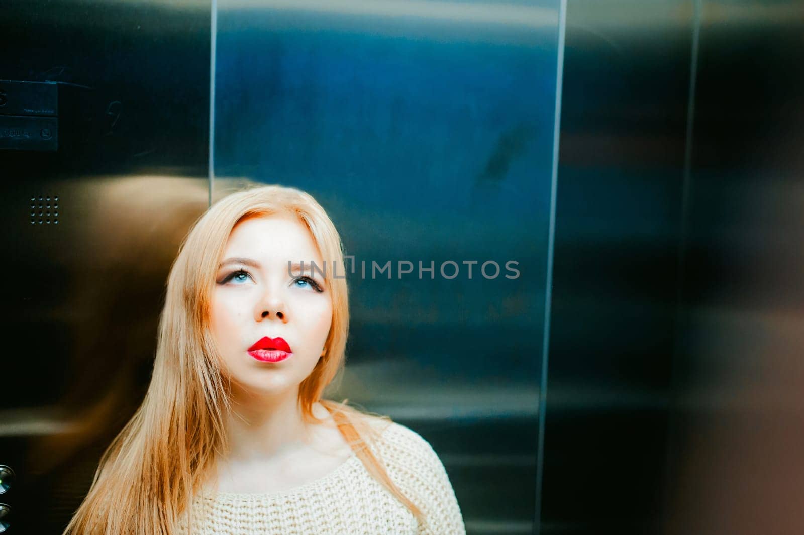 Portrait of a female face, young blond woman with plump lips and beautiful eyes dressed in a knitted sweater. Against elevator lift background