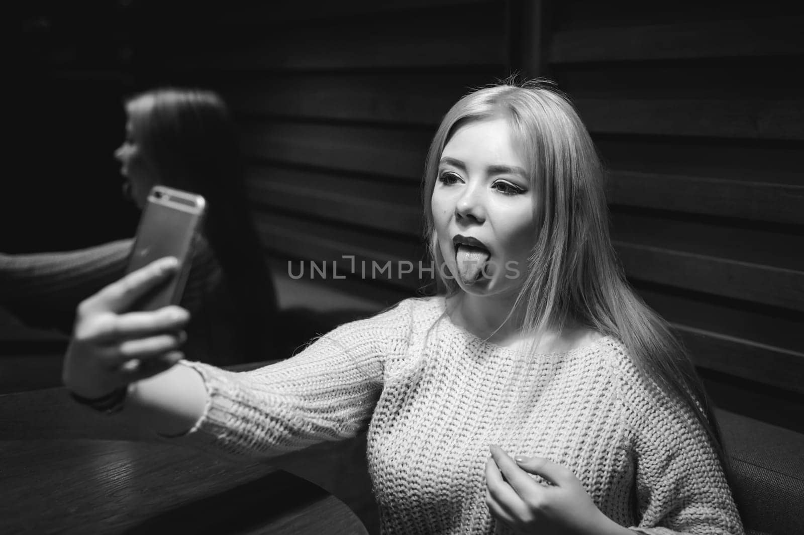 young blond woman in a knitted sweater, making a selfie photo on a smartphone emotionally shows tongue, sitting at a table in a cafe, spending time alone, doing internet communication