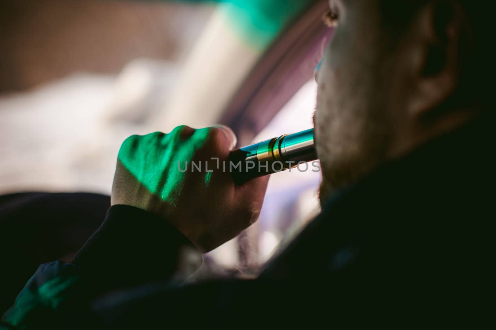young vaper man with beard vaping mechanical mod. Guy smokes an electronic cigarette by blowing a smoke vapor. Bringing rda mouthpiece to the lips on colorful background