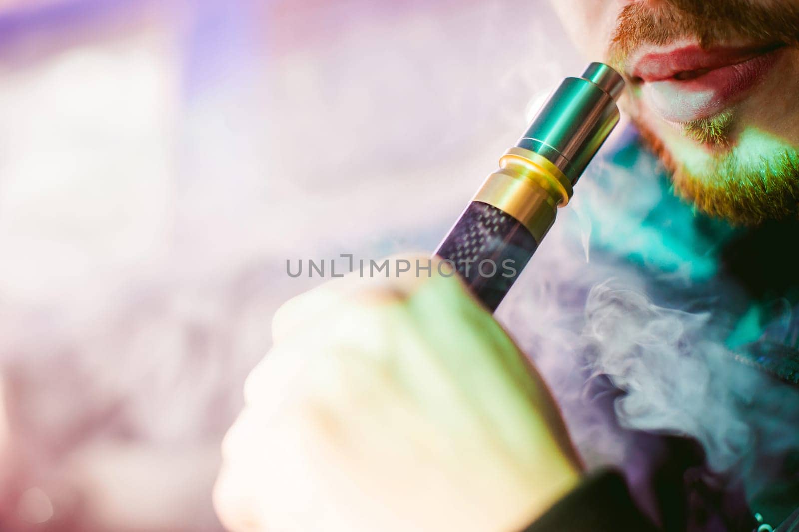 young vaper man with beard vaping mechanical mod. Guy smokes an electronic cigarette by blowing a smoke vapor. Bringing rda mouthpiece to the lips on colorful background