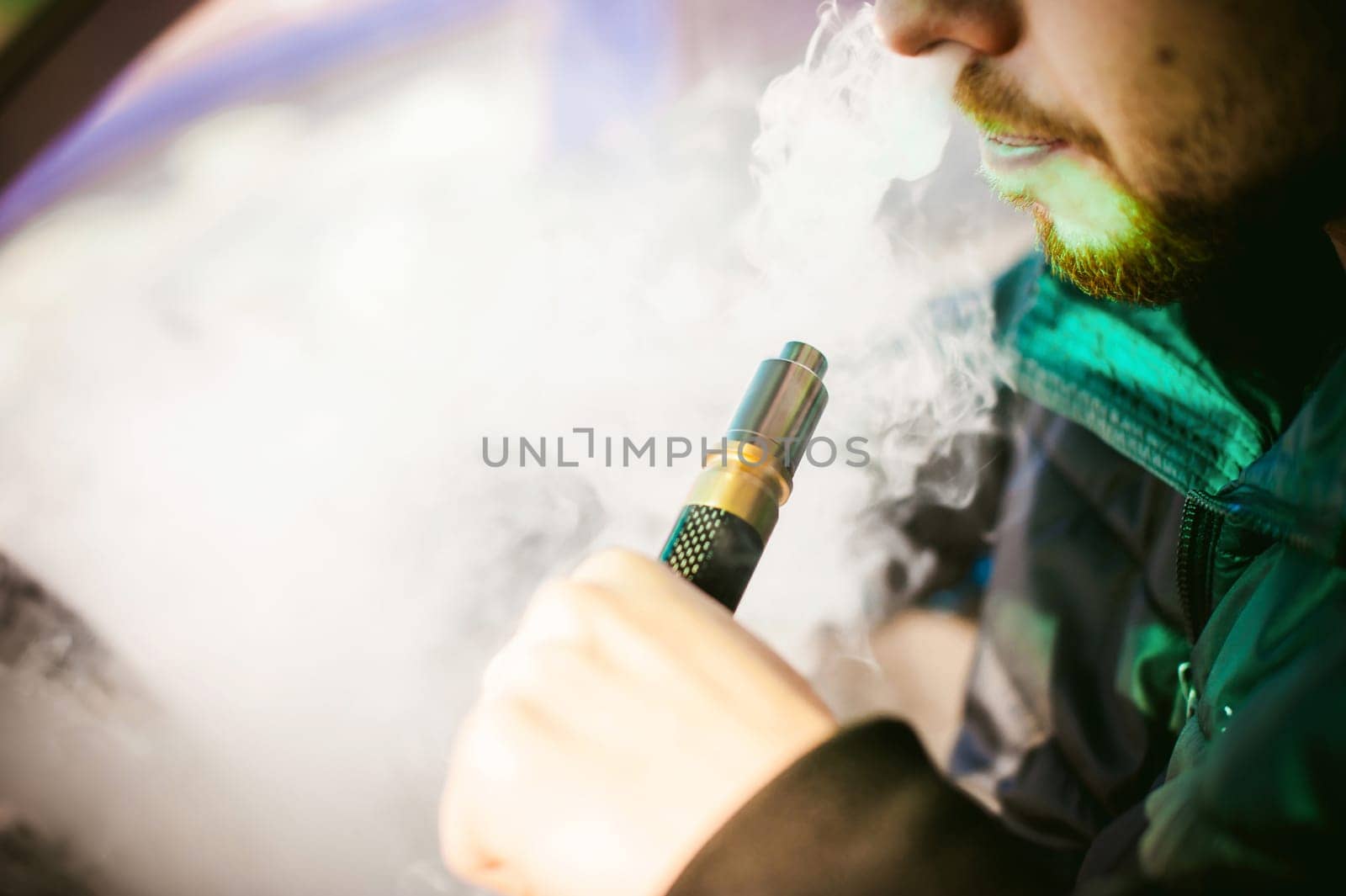 young vaper man with beard vaping mechanical mod. Guy smokes an electronic cigarette by blowing a smoke vapor. Holds in hand on colorful background