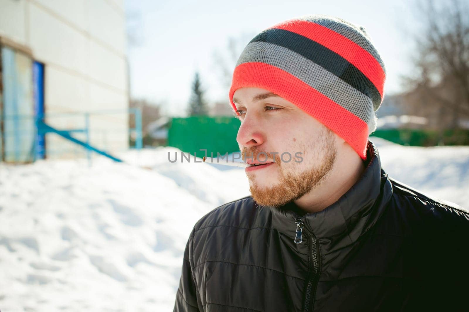 Portrait of a man with a beard with a mustache, smoking a cigarette, with an emotional face in the open air. Dressed in a bright striped hat, against the background of a snowy sunny day