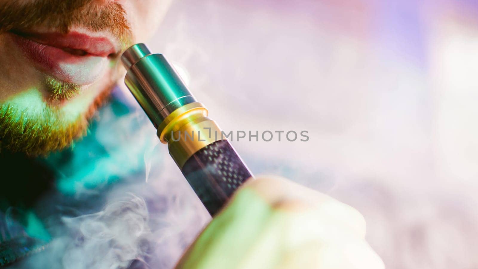 young vaper man with beard vaping mechanical mod. Guy smokes an electronic cigarette by blowing a smoke vapor. Bringing rda mouthpiece to the lips on colorful background