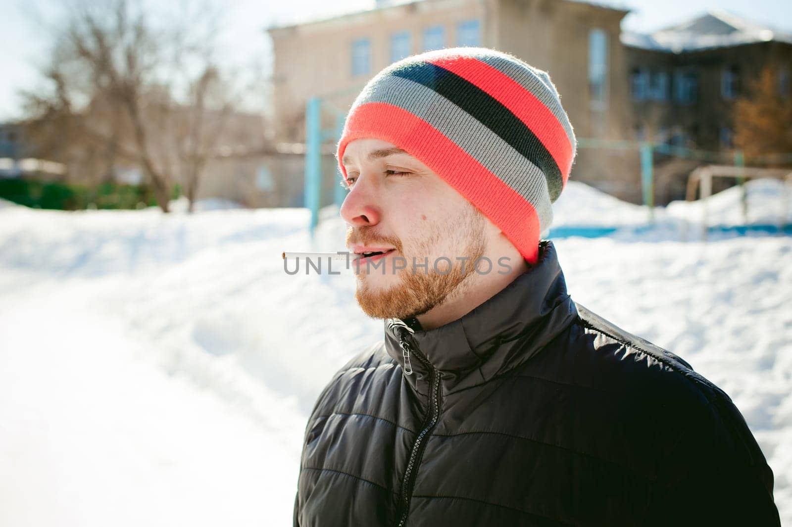 Portrait of a man with a beard with a mustache, smoking a cigarette, with an emotional face in the open air. Dressed in a bright striped hat, against the background of a snowy sunny day