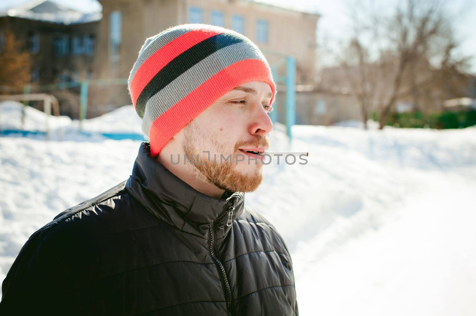 Portrait of a man with a beard with a mustache, smoking a cigarette, with an emotional face in the open air. Dressed in a bright striped hat, against the background of a snowy sunny day