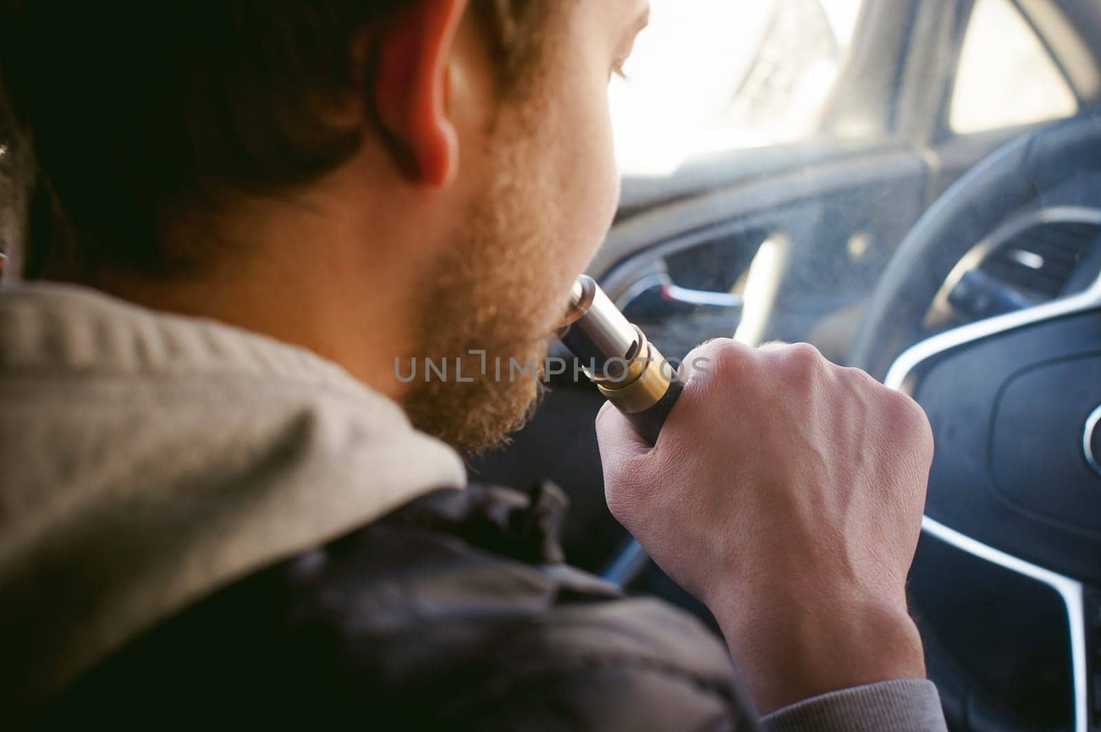 young vaper man with beard vaping mechanical mod. Guy smokes an electronic cigarette by blowing a smoke vapor. Bringing rda mouthpiece to the lips on colorful background Sitting at the wheel of a car
