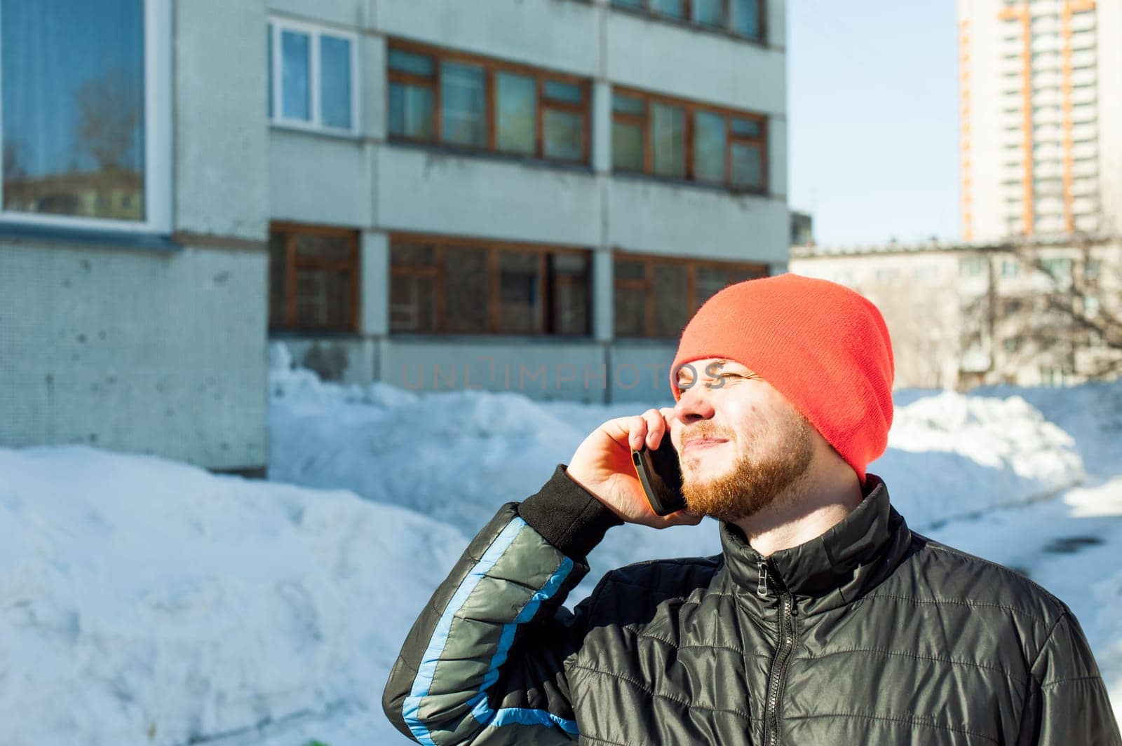 man Emotionally communicates by telephone. guy with beard, dressed in an autumn sports casual jacket and bright orange hat, Speaks by cell phone in stylish case, walking outdoors on sunny winter day