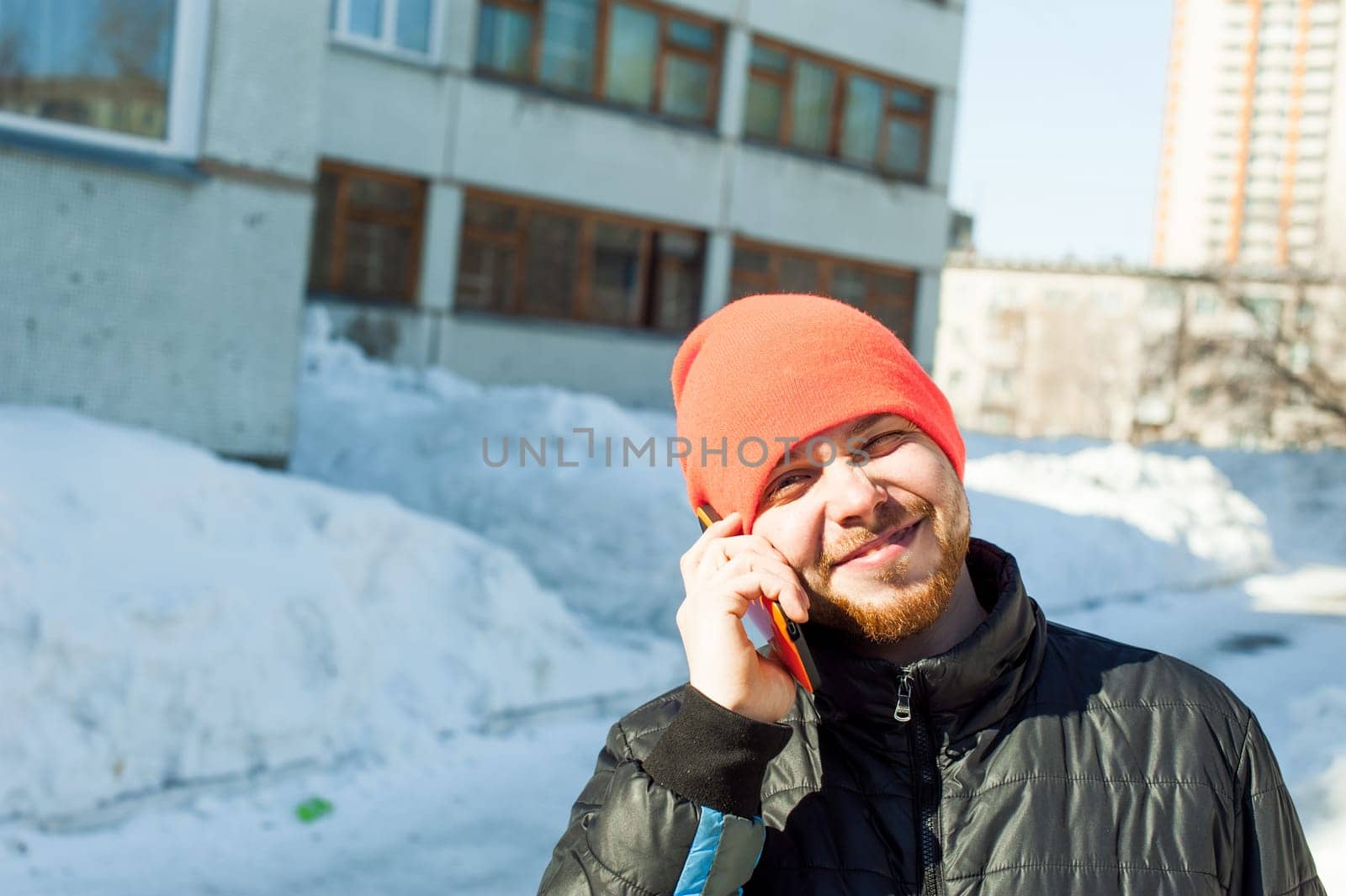 man Emotionally communicates by telephone. guy with beard, dressed in an autumn sports casual jacket and bright orange hat, Speaks by cell phone in stylish case, walking outdoors on sunny winter day