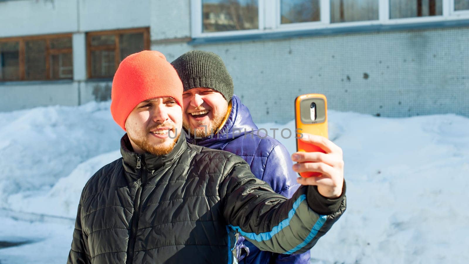 Two men photographing themselves.guy with beard,dressed in autumn sports casual jacket and bright orange hat, makes selfie on phone in stylish case,walking with his friend outdoors sunny winter day