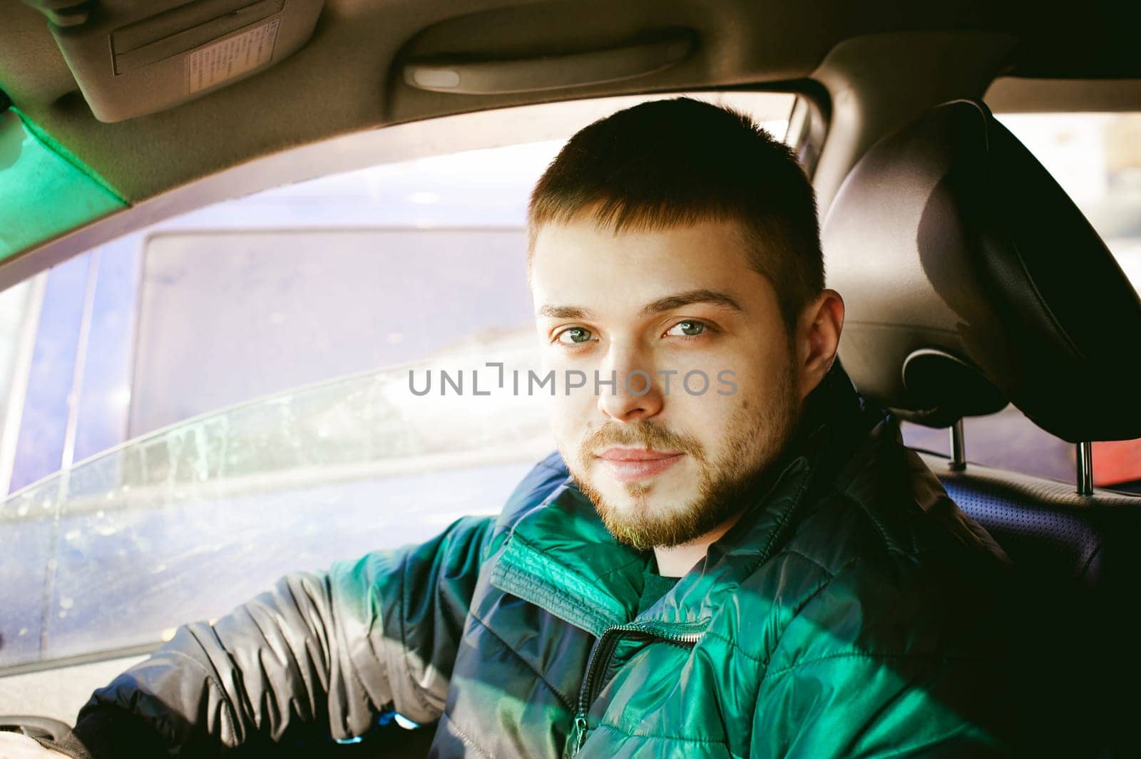 young male driver sits holding his hand behind the wheel in the salon of his car, a happy look of a bearded motorist man