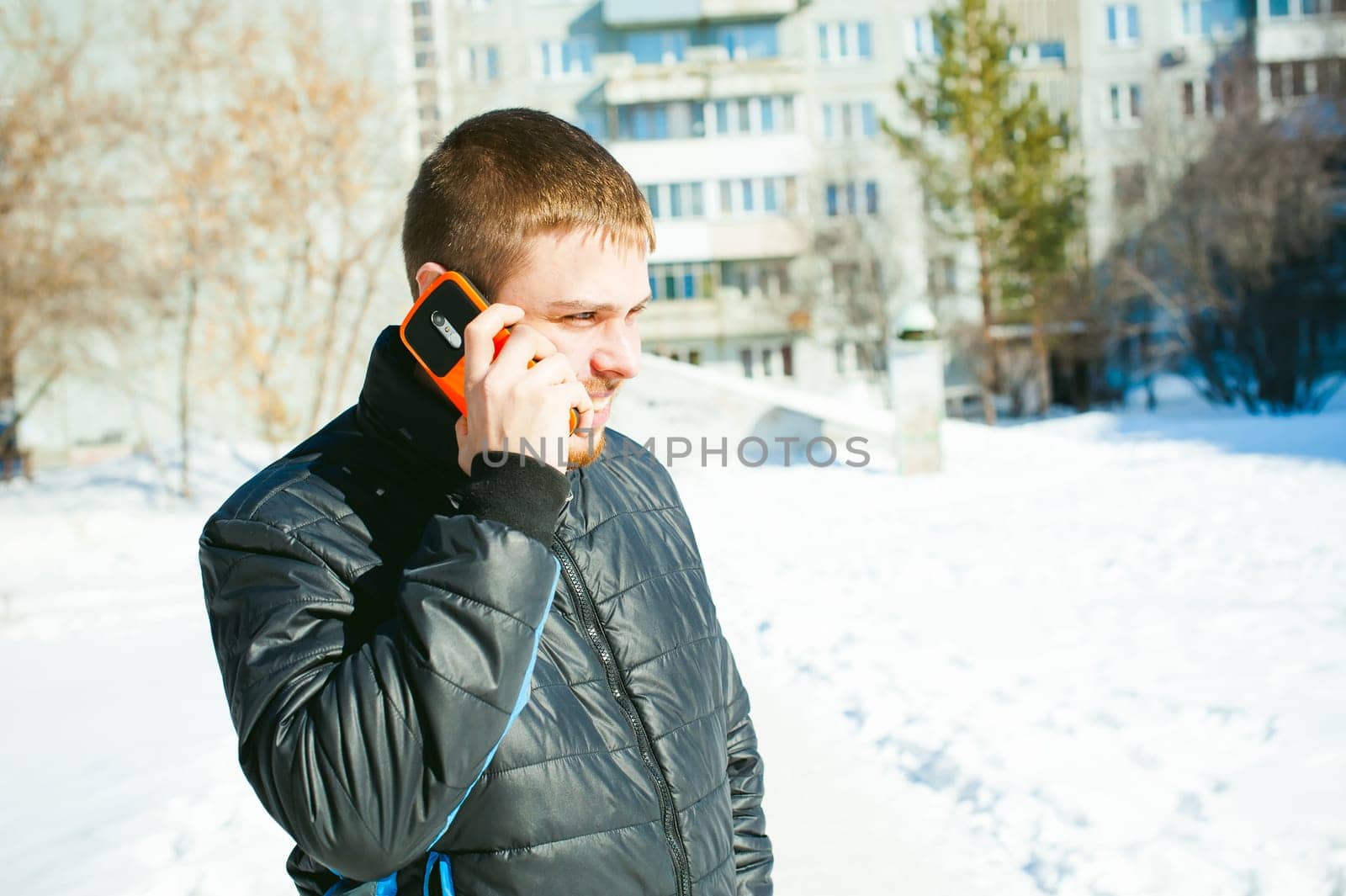 Telephone calls men. The guy is talking on cell phone in street in winter, dressed in warm clothes, holds in his hand phone at ear, in bright orange sheath case