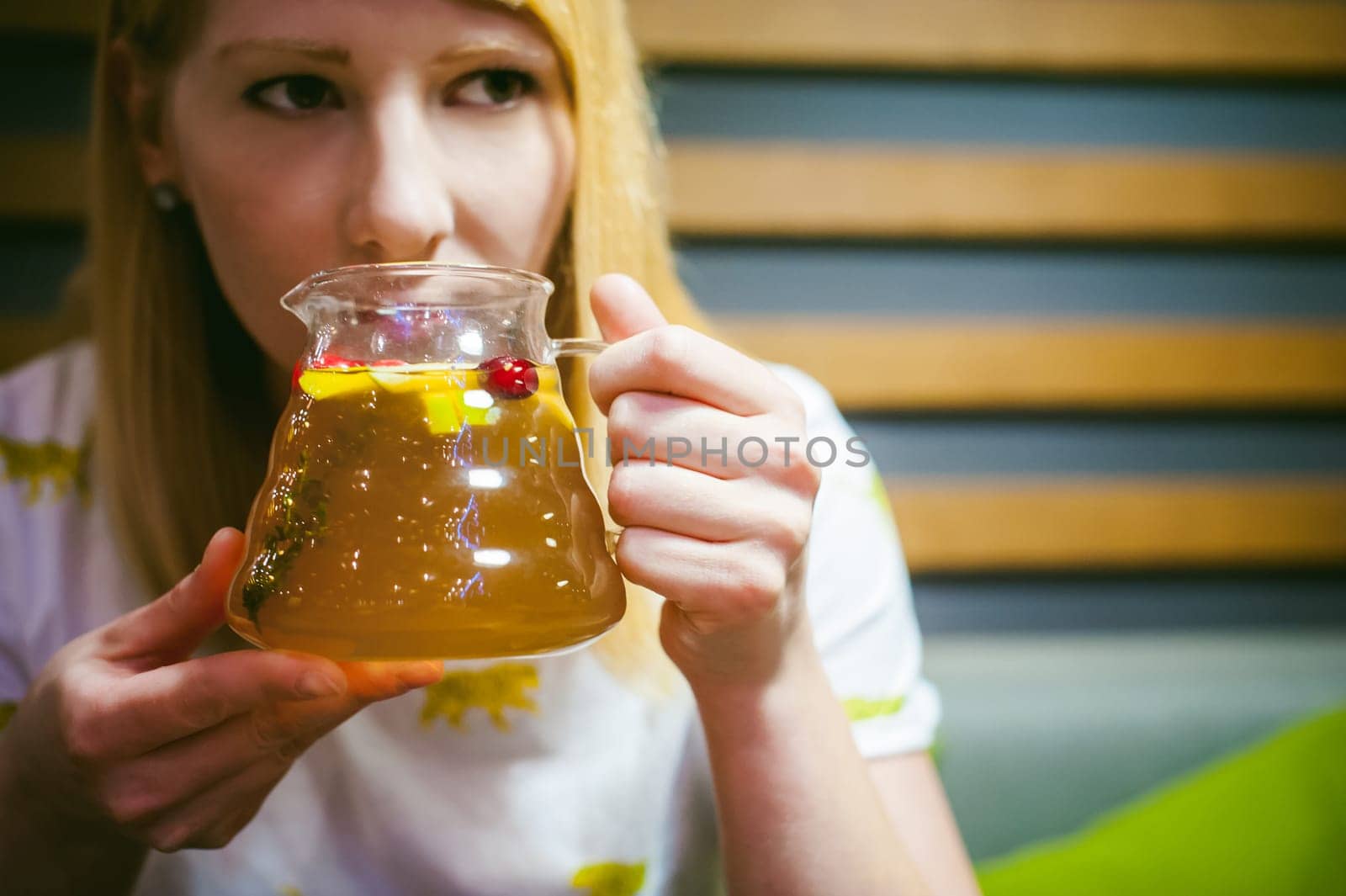Young blonde woman wearing white T-shirt with print, girl Holds a glass jug in his hand, sitting at a table in a cafe, background stylized Wood texture lit up with warm light