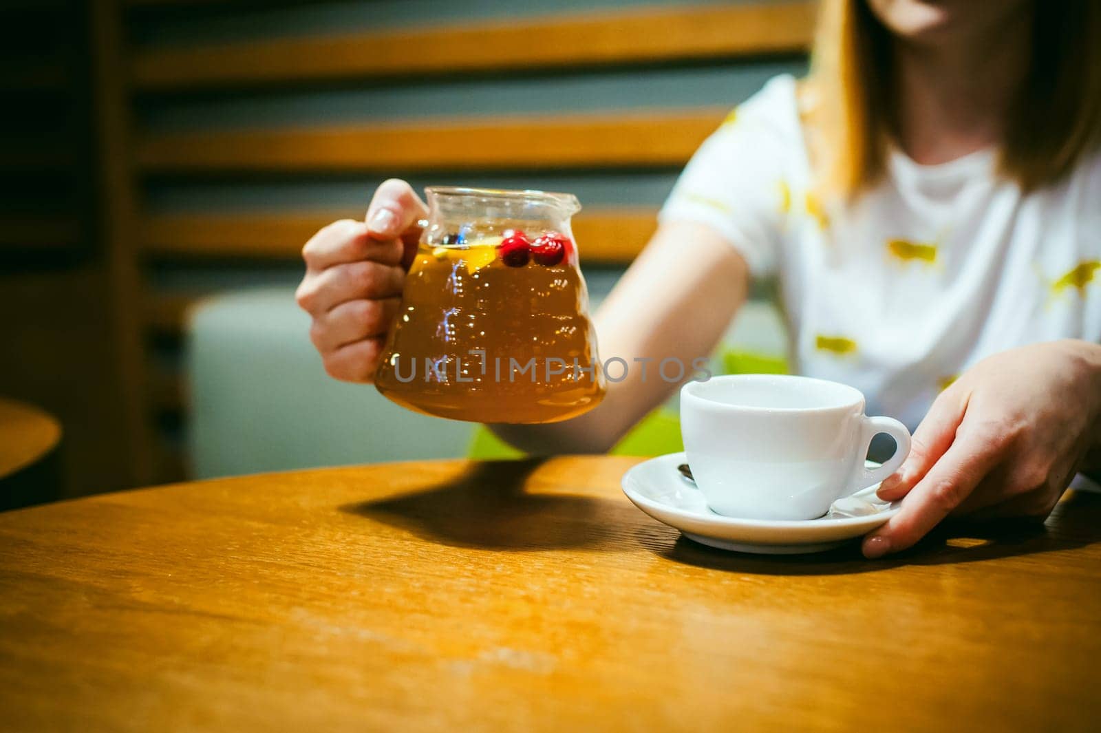 Young blonde woman wearing white T-shirt with print, girl pours a fruit drink from a glass jug, into a white cup, sitting at a table in a cafe, background stylized Wood texture lit up with warm light