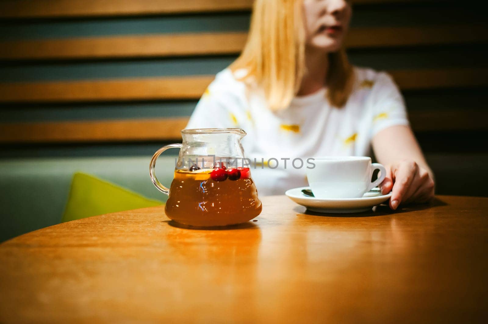 Young blonde woman wearing white T-shirt with print, visited cafe lunch time, drinking fruity hot drink from glass teapot white cup hand holding, background stylized tree table lit up with warm light