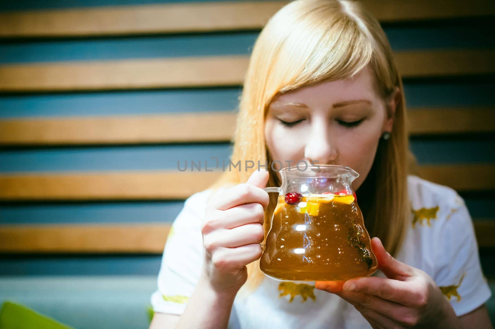 Young blonde woman wearing white T-shirt with print, girl Holds a glass jug in his hand, sitting at a table in a cafe, background stylized Wood texture lit up with warm light