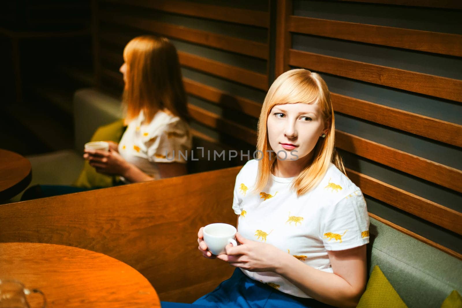 Young blonde woman wearing white T-shirt with print, visited cafe lunch time, drinking fruity hot drink from glass teapot white cup hand holding, background stylized tree table lit up with warm light