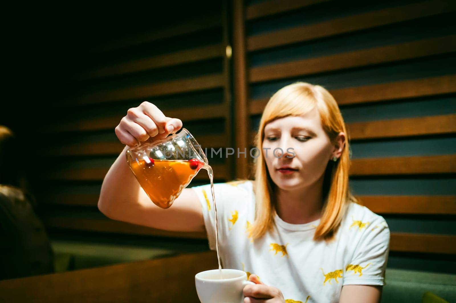 Young blonde woman wearing white T-shirt with print, girl pours a fruit drink from a glass jug, into a white cup, sitting at a table in a cafe, background stylized Wood texture lit up with warm light