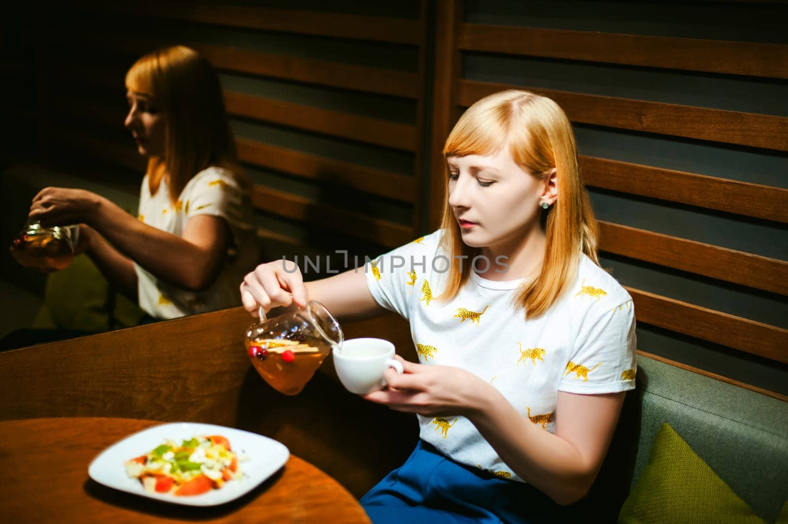 Young blonde woman wearing white T-shirt with print, girl pours a fruit drink from a glass jug, into a white cup, sitting at a table in a cafe, background stylized Wood texture lit up with warm light