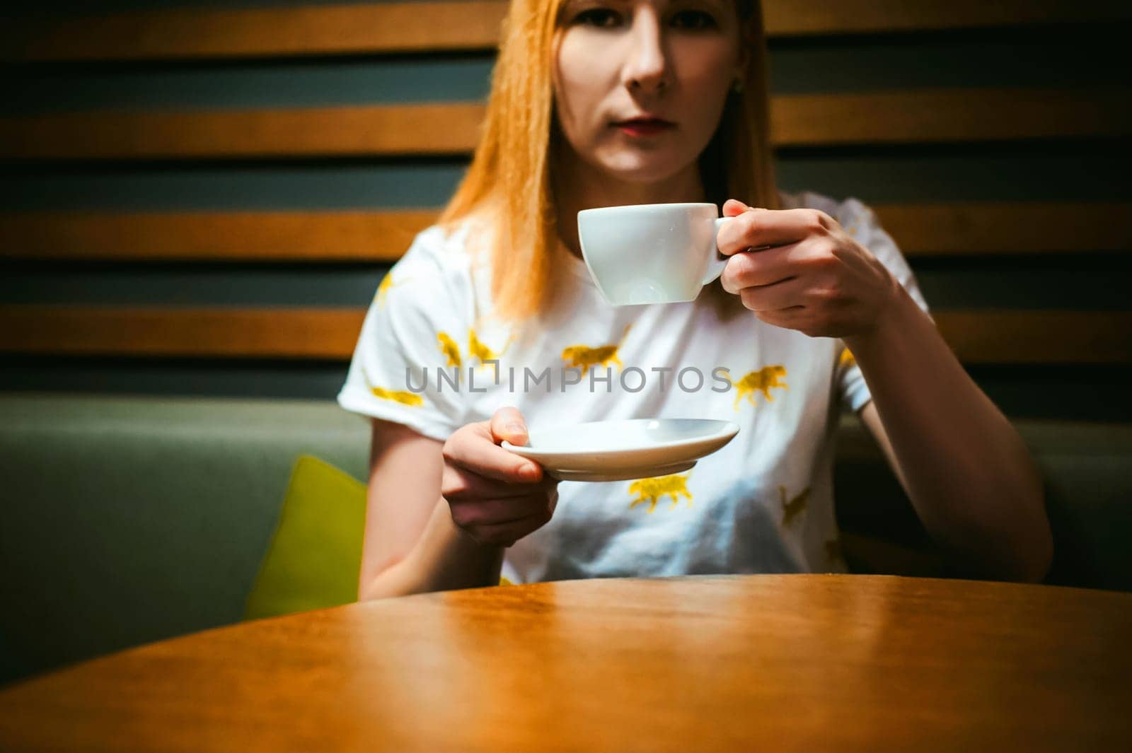 Young blonde woman wearing white T-shirt with print, girl Drinks from a white porcelain cup holding it in his hands, sitting at a table in a cafe, background stylized Wood texture lit up with warm light