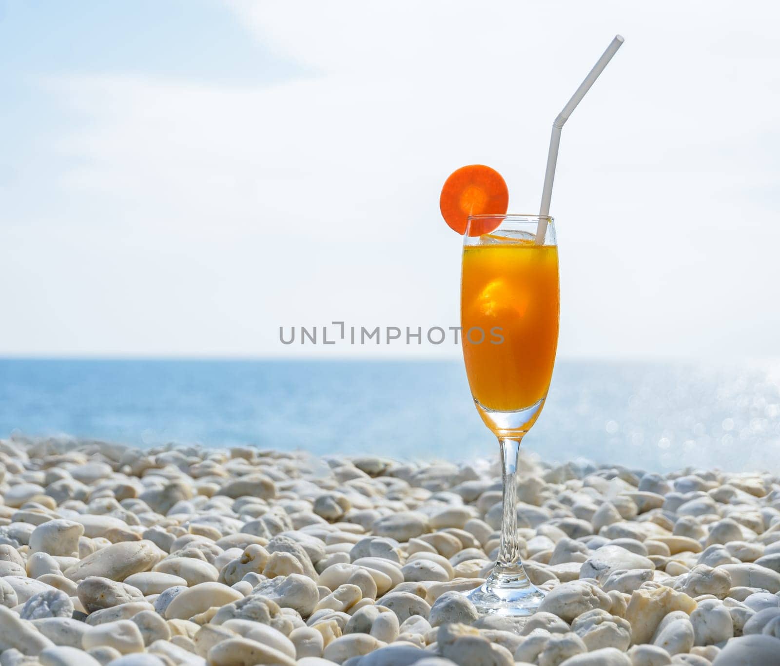 Orange juice with carrot slice in cocktail glass on white pebbly beach with seascape background, summer tropical holiday concept