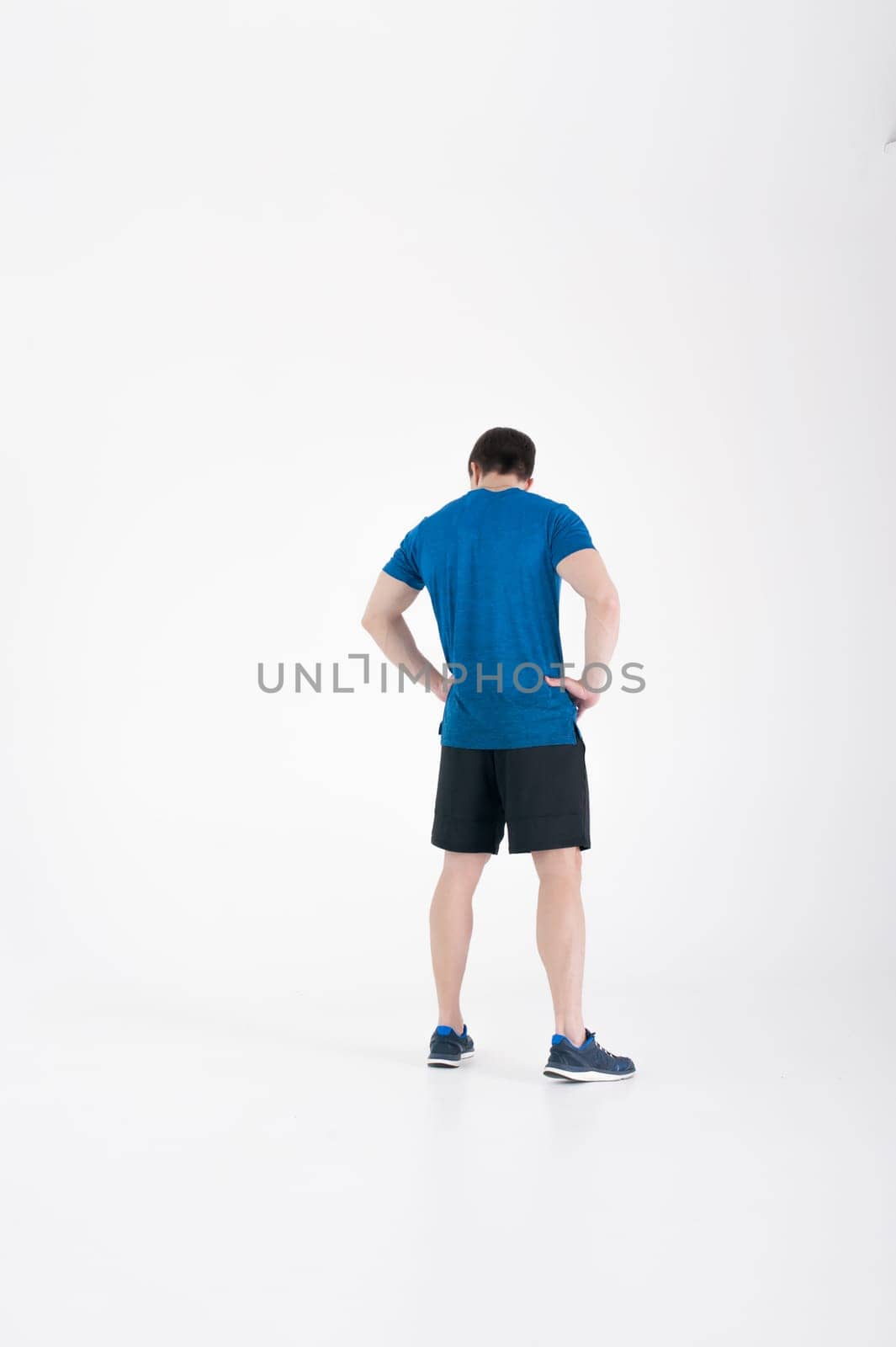Young muscular handsome athletic man dressed in blue sport shirt, shorts and sneakers standing on white background in studio, Holding hands at waist