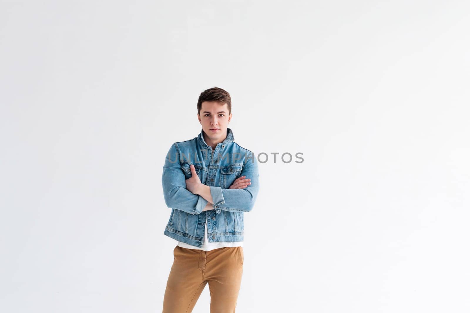 Young handsome man of strong Athlete's body, dressed in Blue jeans jacket in style of old school and white t-shirt with brown trousers, Posing on white studio background, With his arms crossed