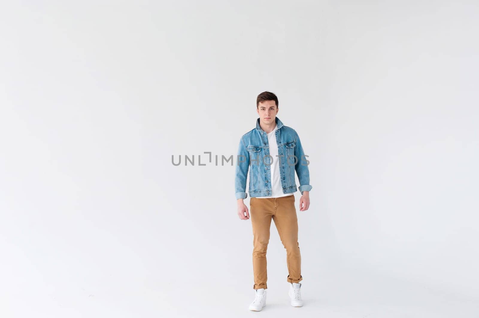 Young handsome man of strong Athlete's body, dressed in Blue jeans jacket in style of old school and white t-shirt with brown trousers, Posing on white studio background