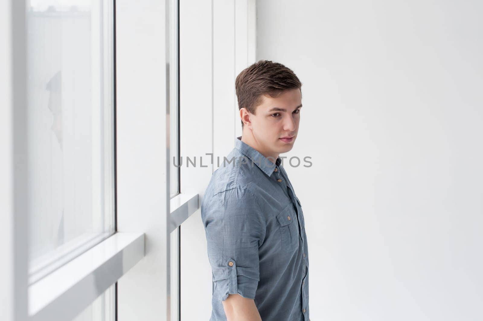 Young handsome man of strong athletic build, dressed in a shirt and brown trousers, Posing on white studio background, near window in full wall, hugging her back